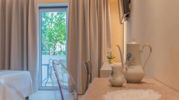 A hotel room with a bed, a table with a vase, a chair, and a view of greenery outside.