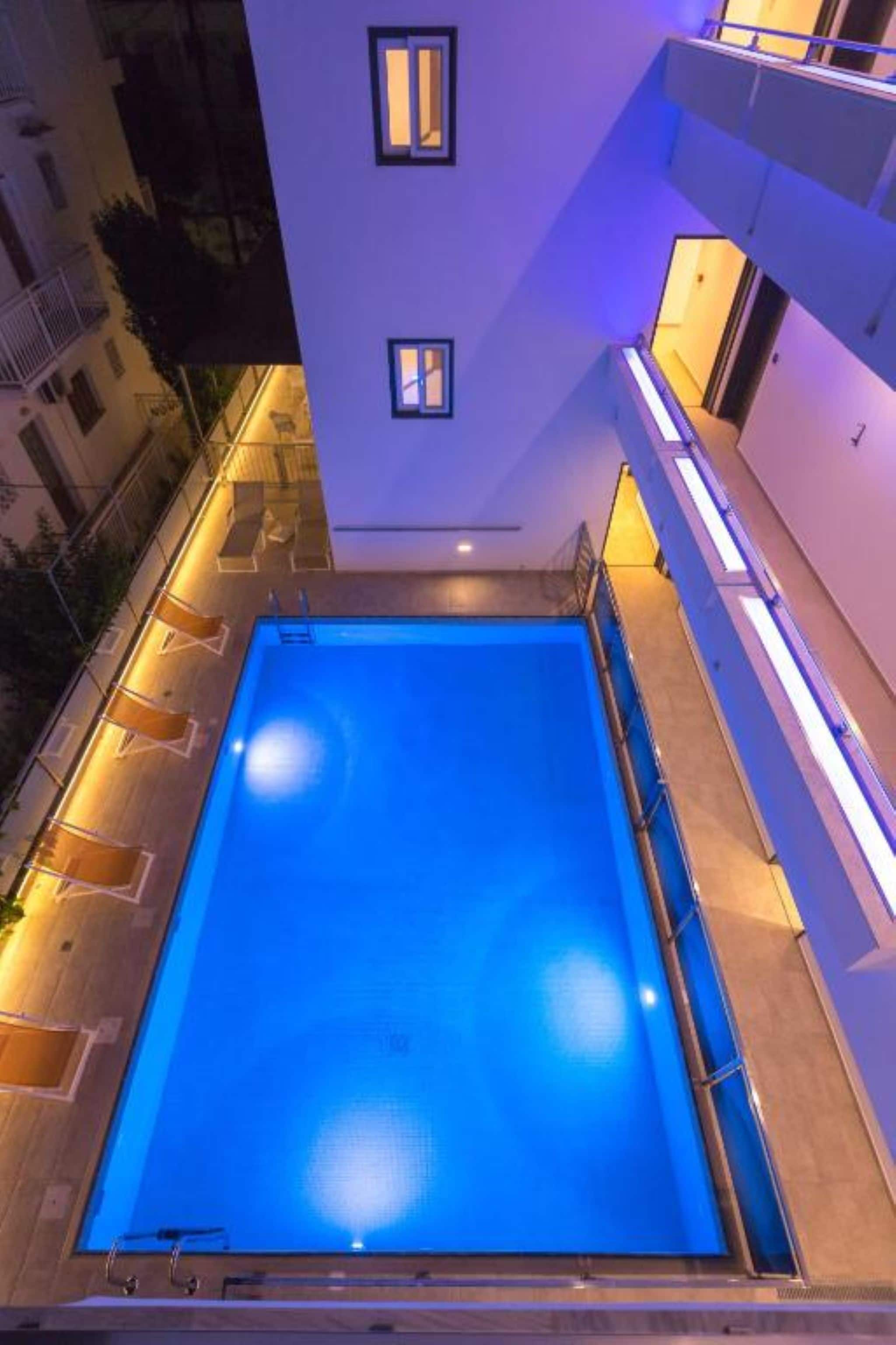 Una piscina en la azotea con superficie de agua azul cristalina, rodeada por un edificio moderno con balcones y fachada bien iluminada.