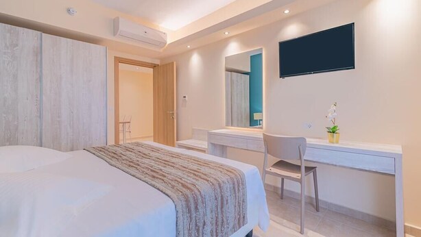 A modern bedroom with a large bed, a desk, and a TV mounted on the wall. - Christa (Kos)