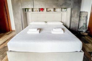 Un lit bien fait, avec du linge de lit blanc et deux serviettes, une tête de lit grise et une étagère au-dessus, ornée de petits objets décoratifs.