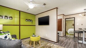 A living room with a green accent wall, a flat-screen TV, a ceiling fan, a small yellow ottoman, and a grey sofa.