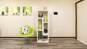 A room with a green chair, a white shelving unit with decorative items, and three paintings on the wall.