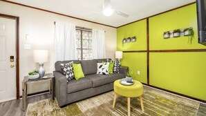 A living room with a grey sofa, a yellow ottoman, a ceiling fan, and a window with white curtains.