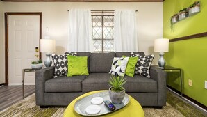 A living room with a grey sofa, green and black patterned cushions, a potted plant, and a tray with remote controls.