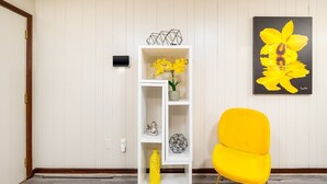 Ein Zimmer mit weißer Wand, einem gelben Sessel, einer weißen Regaleinheit mit Dekoration und einem gerahmten Gemälde einer gelben Blume.