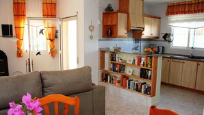 Un salon avec un canapé et des fauteuils, une table à manger agrémentée d’un vase de fleurs, et une cuisine avec des meubles en bois et une bibliothèque.