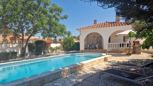 Une piscine aux bords recouverts de pierres, une terrasse avec des chaises et un bâtiment orné d’arches.