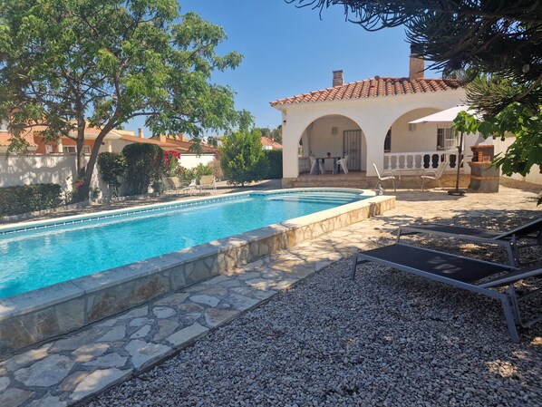 Ein Pool mit Steinfliesen, eine Terrasse mit Stühlen und ein Gebäude mit Bögen.