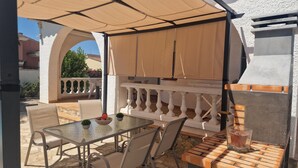Une terrasse extérieure couverte, avec une table et des chaises, un barbecue et une balustrade blanche.