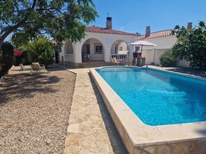 A swimming pool with a stone deck, a patio area with chairs, and a building with arches and a tiled roof.