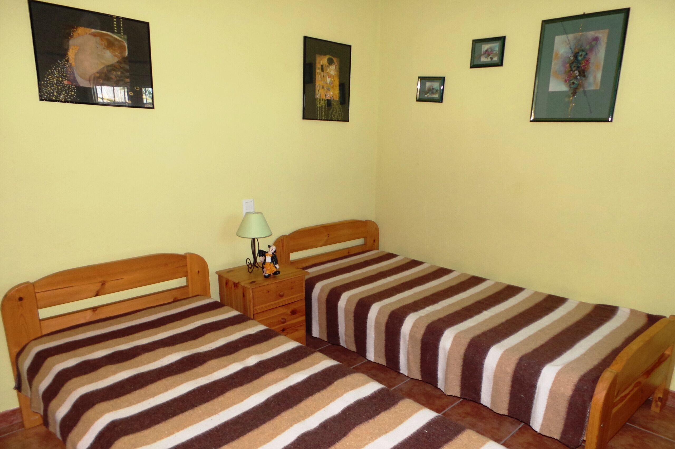 Une chambre avec deux lits simples, une table de chevet en bois avec une lampe, et trois tableaux encadrés accrochés au mur.