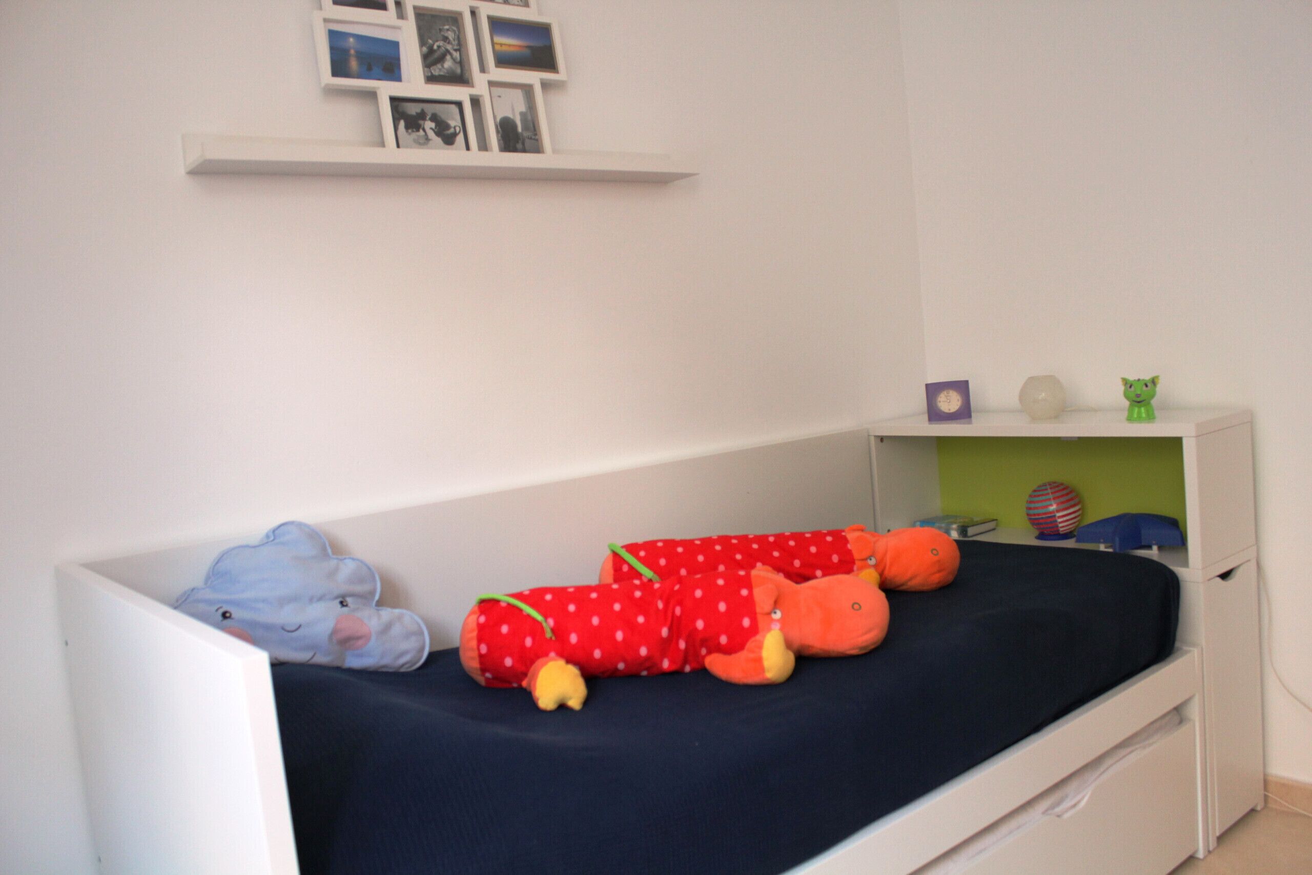 A child's bed with a dark blue mattress, a white headboard, and a white wall with a shelf above.
