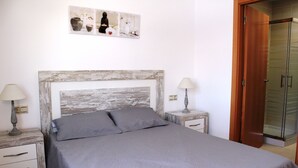 Une chambre avec une tête de lit en bois, deux tables de chevet avec des lampes, un lit avec une literie grise et une porte de douche en verre.