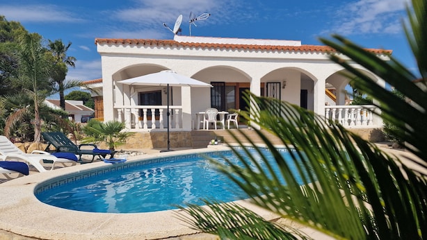 Uma área na beira da piscina com espreguiçadeiras, piscina e um edifício com arcos e antenas parabólicas.