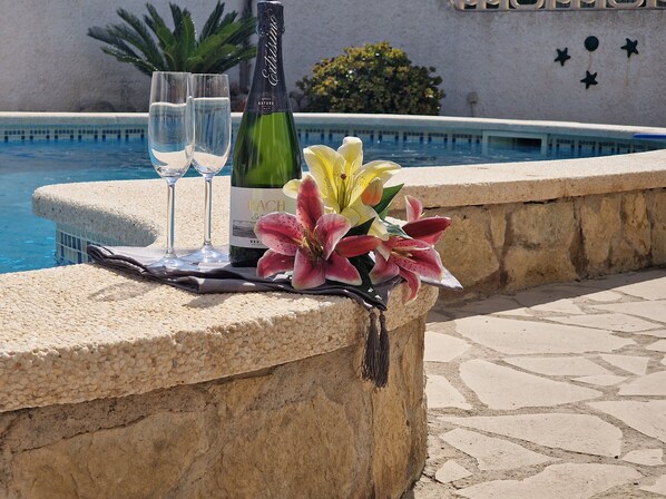 Une table placée au bord de la piscine, avec une bouteille de vin, deux verres et un arrangement floral.