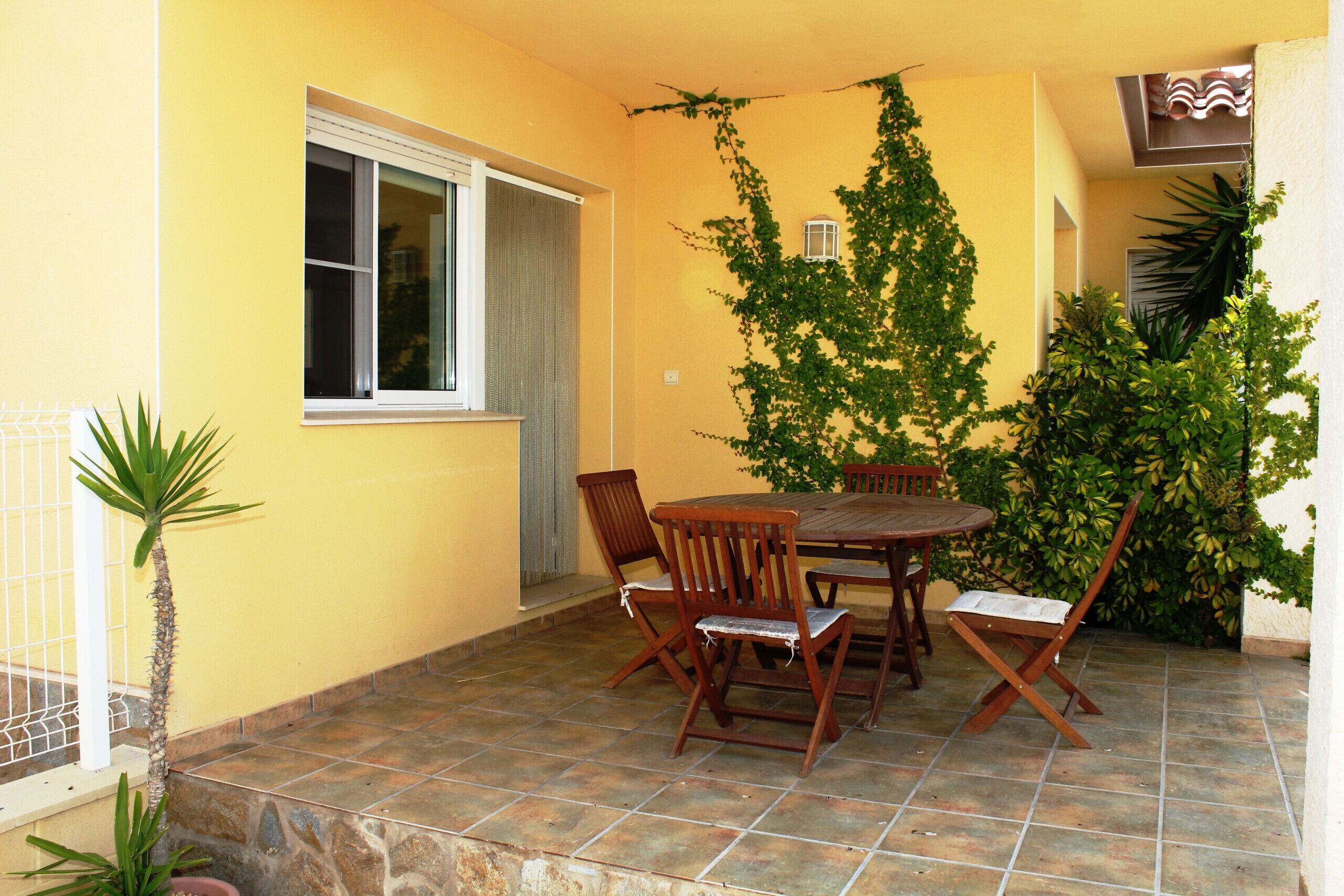 Une terrasse avec une table et des chaises, une fenêtre et un mur recouvert de plantes grimpantes.