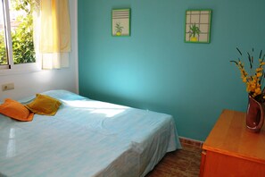 Een kamer met een bed, een raam met gordijnen, een muur met twee ingelijste foto's en een nachtkastje van hout met een vaas.
