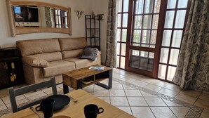 Ein Wohnzimmer mit einer Couch, einem Couchtisch, einem Esstisch mit Tellern und Tassen, einem Spiegel und einem großen Fenster mit Vorhängen.