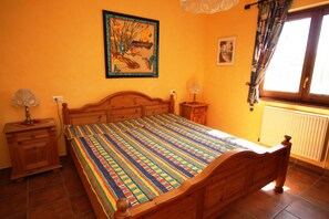 Een slaapkamer met een houten bed, een kleurrijk gestreept dekbed, een nachtkastje met een lamp, een aan de muur hangend schilderij en een raam met gordijnen.