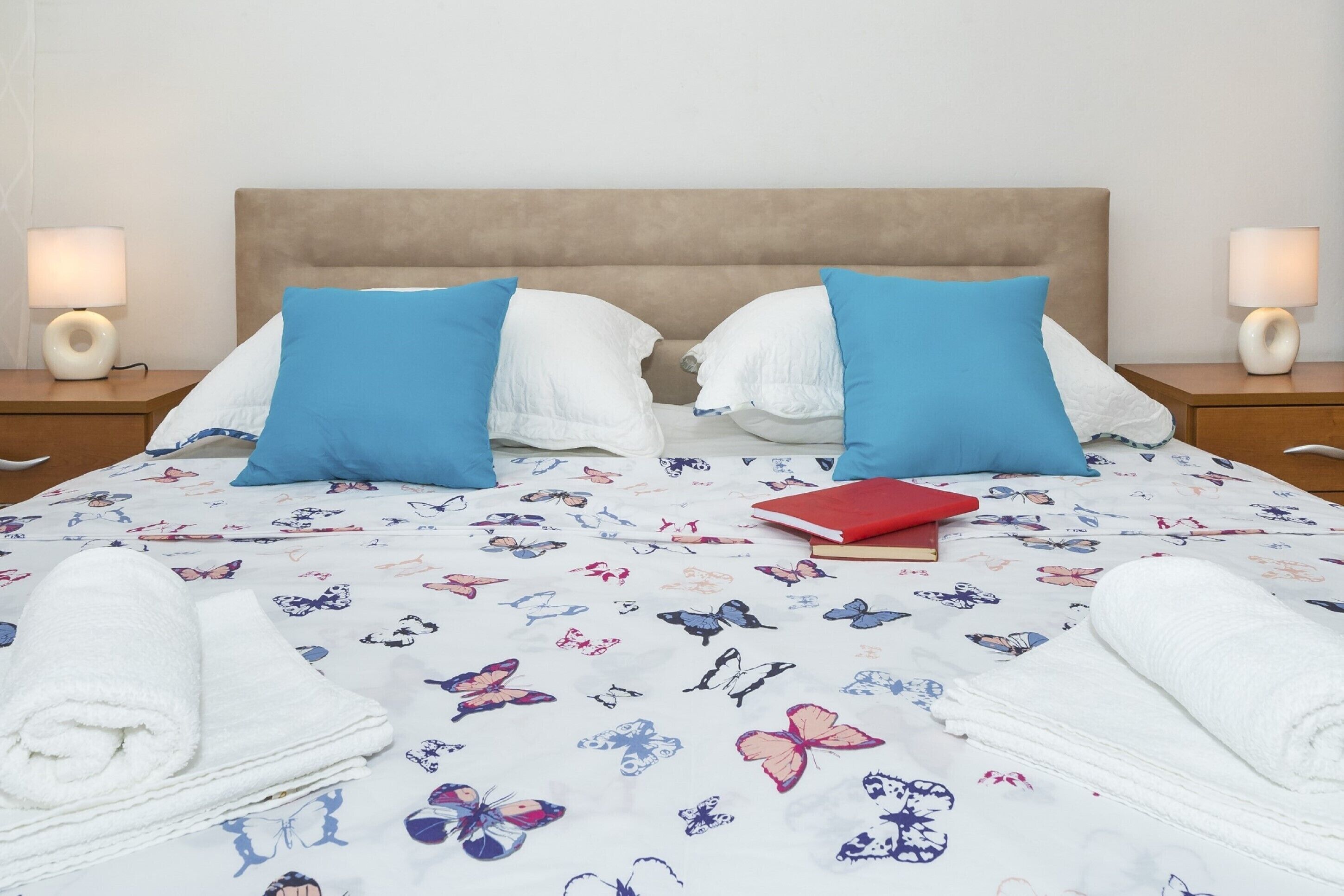 A neatly made bed with a floral-patterned bedspread, two blue pillows, and two white pillows. There are two bedside lamps on either side of the bed.