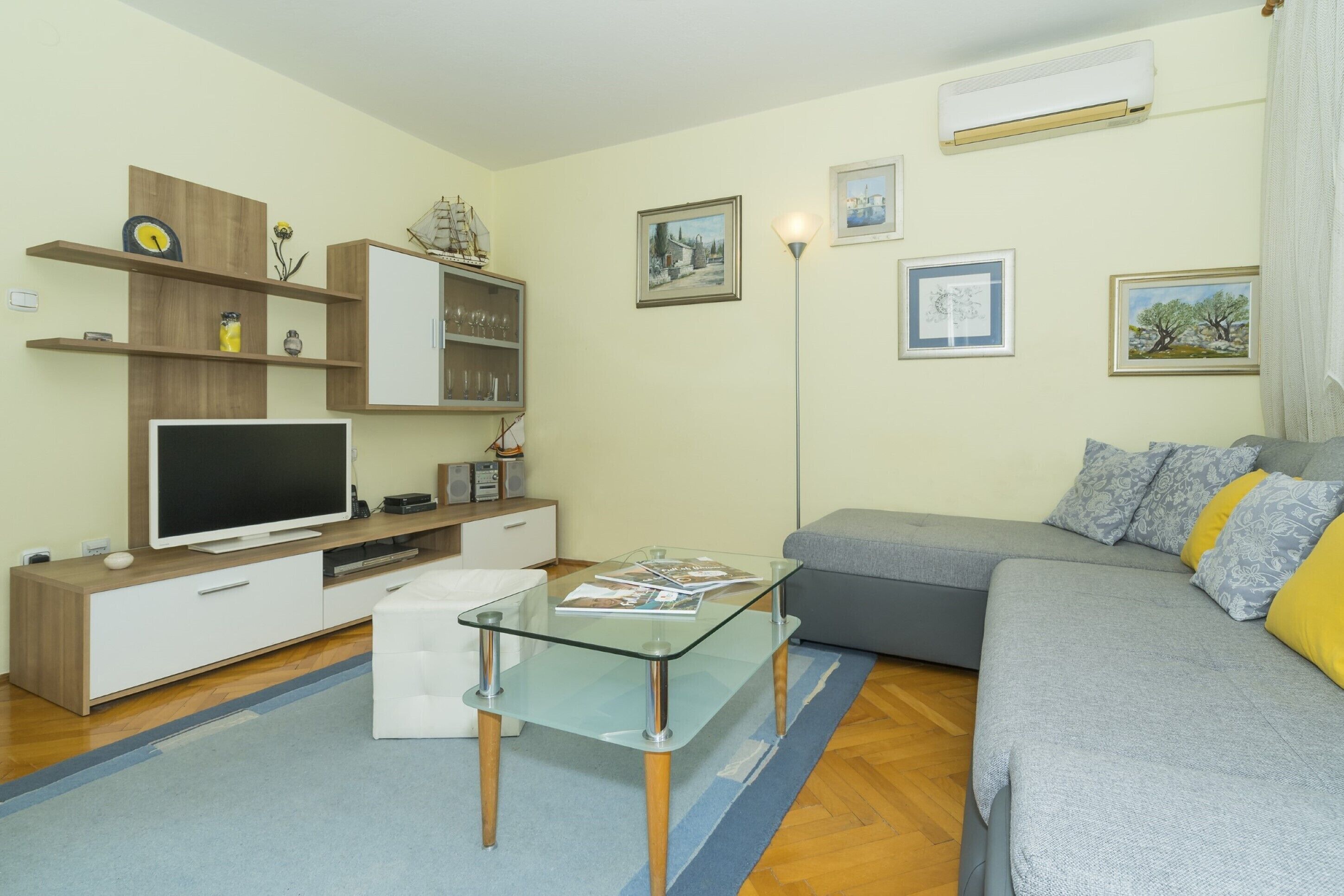 A modern living room with a flat-screen TV, glass coffee table, grey sofa, wooden shelves, and framed pictures on the wall.