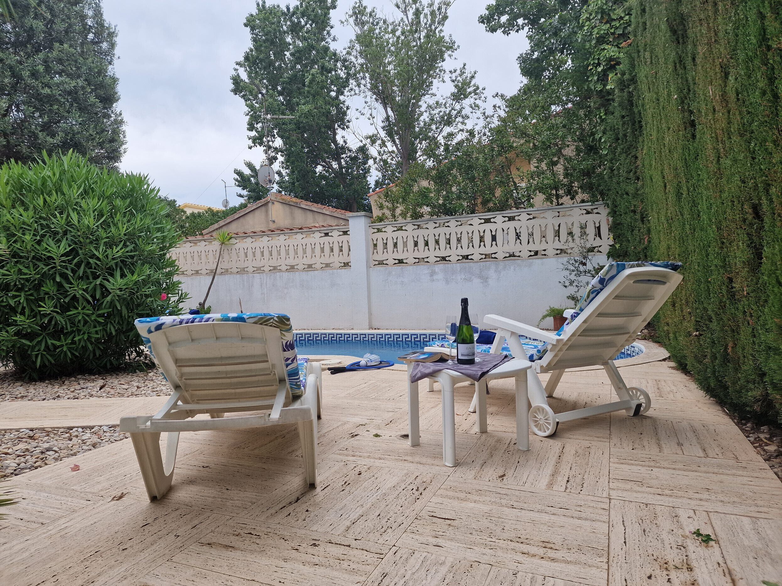Un área junto a la piscina con dos tumbonas, una mesa con una botella y un vaso, y un muro decorativo.