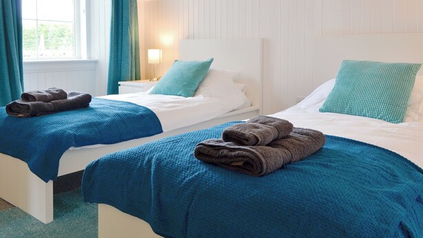 Habitación de hotel con dos camas, cada una con una manta azul y una toalla marrón. Se observa una ventana con cortinas, una lámpara de noche y un cuadro en la pared.