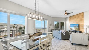 A modern living room with a dining area, large windows, a ceiling fan, and a TV on a wooden wall. - Crystal View 203 3 bedroom/  3 bath unit Beach service great view SO MUCH SPACE (Miramar Beach)