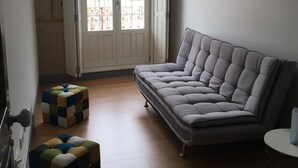 A living room with a grey sofa, a colorful ottoman, and a window with a view of buildings.