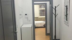 A hallway leading to a bedroom with a bed, a wardrobe, a small fridge, and a tripod.