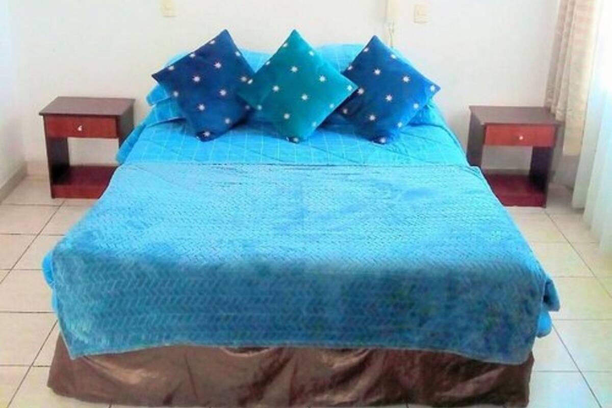 A bed with a blue bedspread and multiple pillows, two bedside tables, and a wall-mounted light fixture.