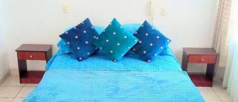 A bed with a blue bedspread and multiple pillows, two bedside tables, and a wall-mounted light fixture.