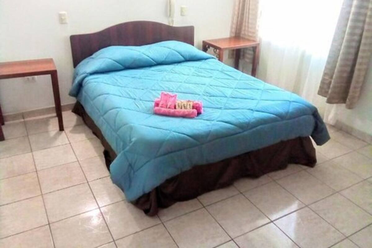 A bed with a blue bedspread and a pink towel, a wooden nightstand, a wooden table, and a white wall with a framed picture.