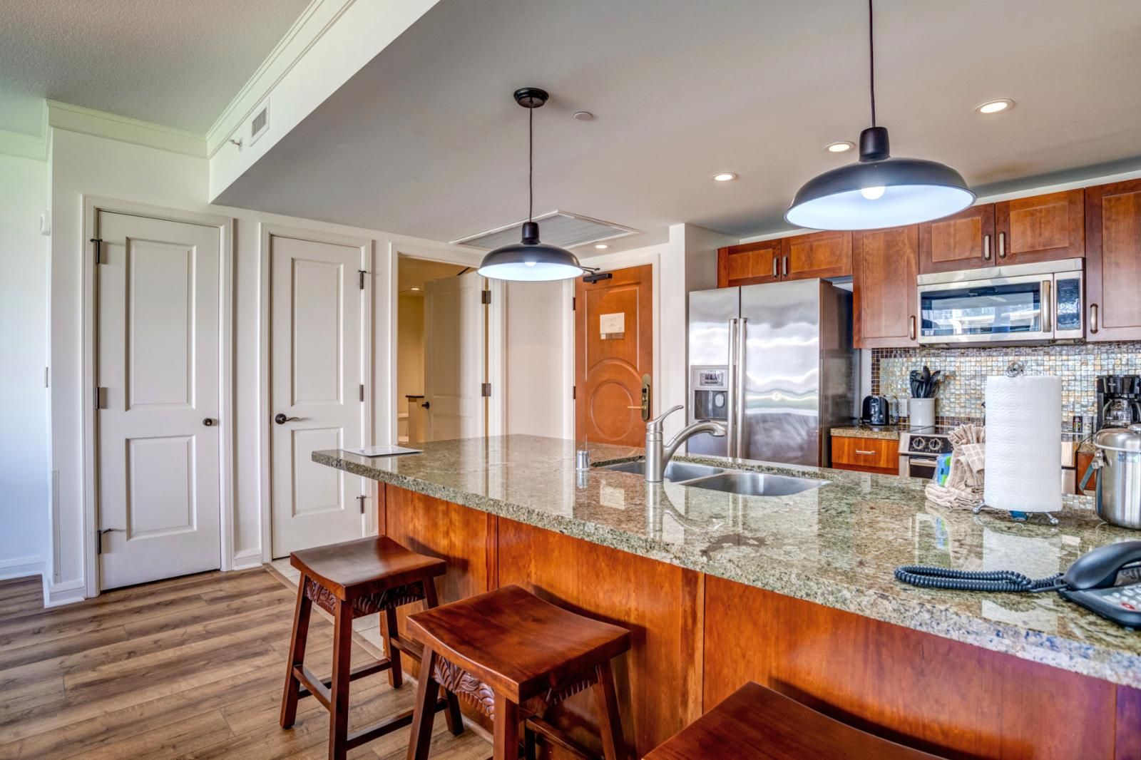 A kitchen with granite countertops, wooden cabinets, a stainless steel refrigerator, and a microwave. There are two bar stools and a ceiling light fixture.