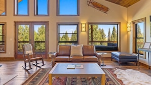 A spacious living room with a leather sofa, a rocking chair, and a coffee table. Large windows offer views of trees and a balcony.