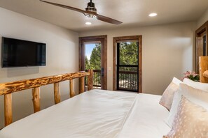 A bedroom with a bed, wooden headboard, a ceiling fan, a TV, and a balcony with a view of trees.