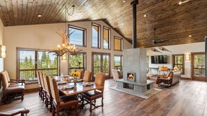 A spacious living room with a large fireplace, wooden flooring, and large windows offering a view of the outdoors.