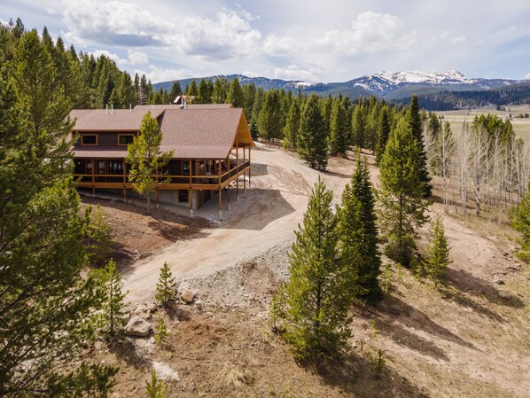 A two-story wooden cabin with a large deck, surrounded by a forest and mountains in the background.