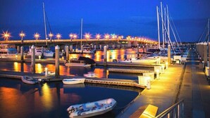 Un port de plaisance au crépuscule, avec des bateaux amarrés, un pont en arrière-plan et des lumières qui illuminent les lieux.
