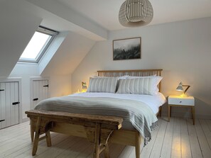 A bedroom with a sloped ceiling, a skylight, a bed with white bedding, a wooden bench, a bedside table with a lamp, and a painting on the wall. - Lower Hilton Cottage (Culrain, Lairg)