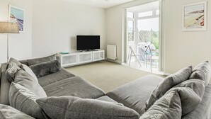 A modern living room with a grey sectional sofa, a flat-screen TV on a white console, and a large sliding glass door leading to a balcony.