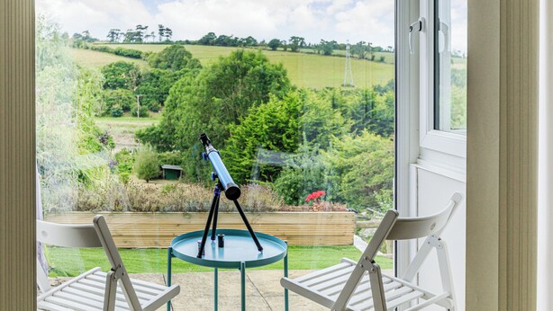 A telescope on a tripod outside a window overlooking a green landscape.