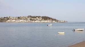 A calm waterfront with several boats, a sandy beach, and a town with buildings on a hill in the background.
