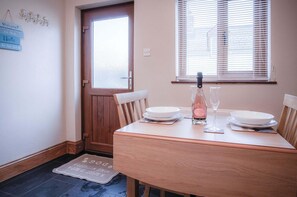 Ein Essbereich mit einem Holztisch, gedeckt für zwei Personen, einer Flasche Roséwein und einem Glas. Eine Tür mit dem Schild 'Welcome to Our Home by the Sea'.