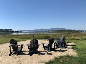 Une plage avec quatre chaises Adirondack disposées sur le sable, offrant une vue sur un lac paisible et des montagnes lointaines.