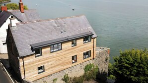 Een modern huis met twee verdiepingen, een stenen basis en houten bovenverdieping, met uitzicht op een rustige zee met boten en verre bergen.