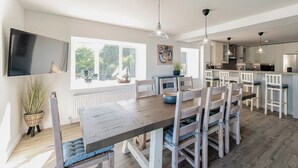 Een moderne keuken met een eethoek, met een houten tafel, witte stoelen, hanglampen en een groot raam met uitzicht op groen.