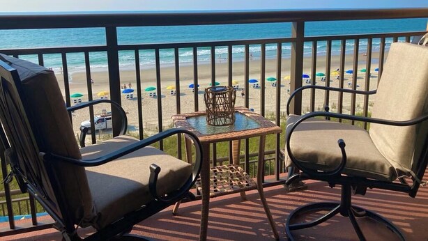Un balcon avec deux chaises et une petite table, donnant sur une plage parsemée de parasols et où se trouvent des gens.
