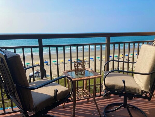 A balcony with two chairs and a small table, overlooking a beach with umbrellas and people.