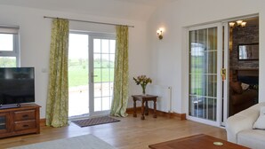 A spacious living room with a wooden floor, a television, a coffee table, and a sofa. There is a sliding glass door leading to a balcony with a view of a green landscape.